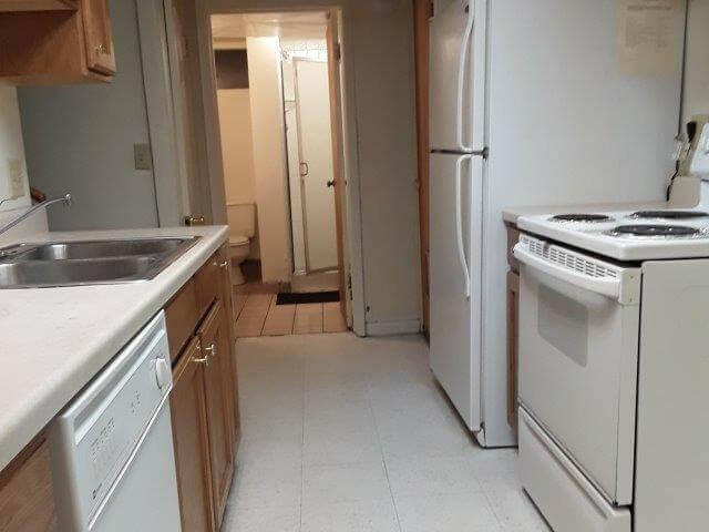 Kitchen showing oak cabinets, off-white countertops, dishwasher, oven, refrigerator, and door to bathroom with bathroom beyond.