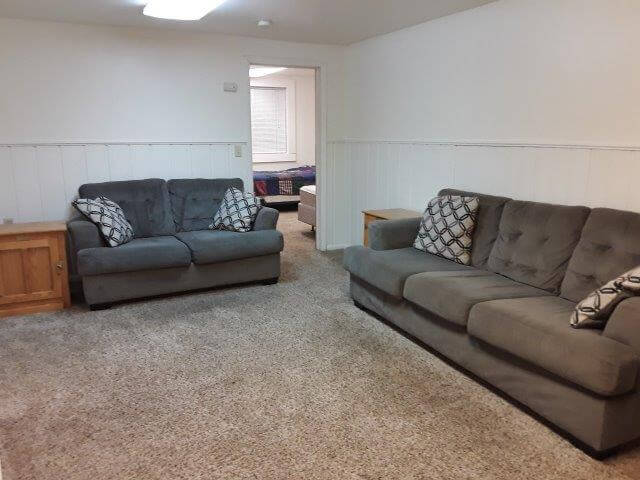 Apt #2 living room with grey sofa/loveseat, oak end tables, off-white walls, and beige carpet; door opens into bedroom beyond.