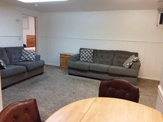 Apt #2 living room with grey sofa/loveseat, oak end tables, off-white walls, and beige carpet; door opens into bedroom beyond.